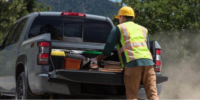 Person wearing a high visibility vest and hard had loading gear into the bed of the 2024 Nissan Frontier to demonstrate dampended assist tailgate