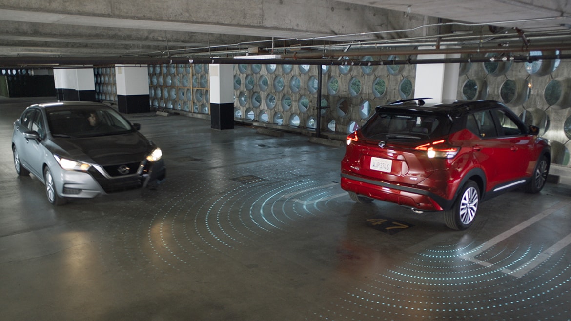 Nissan Kicks rouge 2024 dans une aire de stationnement avec un deuxième véhicule, illustrant les caractéristiques du bouclier de sécurité 360 et de la caméra de recul