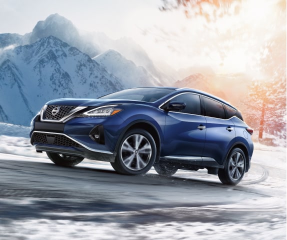 A blue 2024 Nissan Murano driving up a snow covered road with a mountain and trees in the background