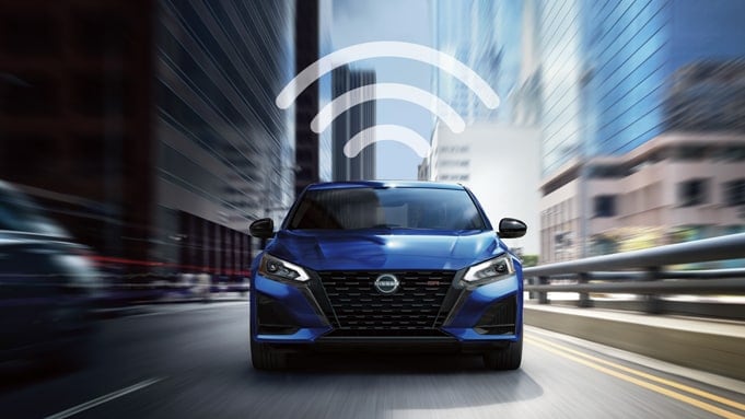 Front view of a blue 2024 Nissan Altima driving with a blurry background and a 3 curved line wifi logo over it