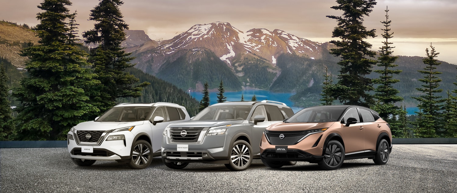 Nissan Rogue and Nissan Pathfinder parked side ny side across from trees and rocky mountains.