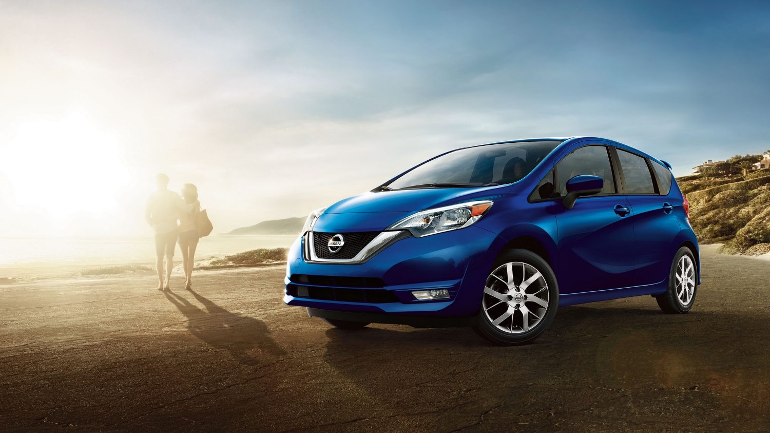 A couple walks away from their blue Nissan Versa Note hatchback parked on a beach