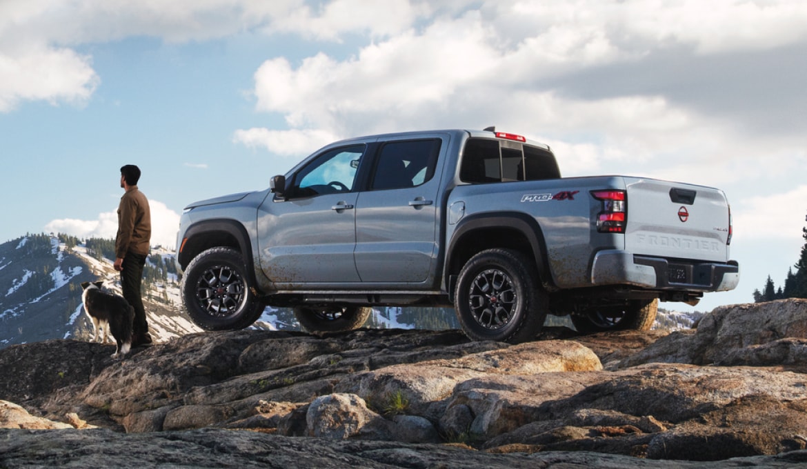 Grey 2024 Nissan Frontier parked on a hill. A person stands in front of the vehicle, looking out at the view. A mountain rises in the background