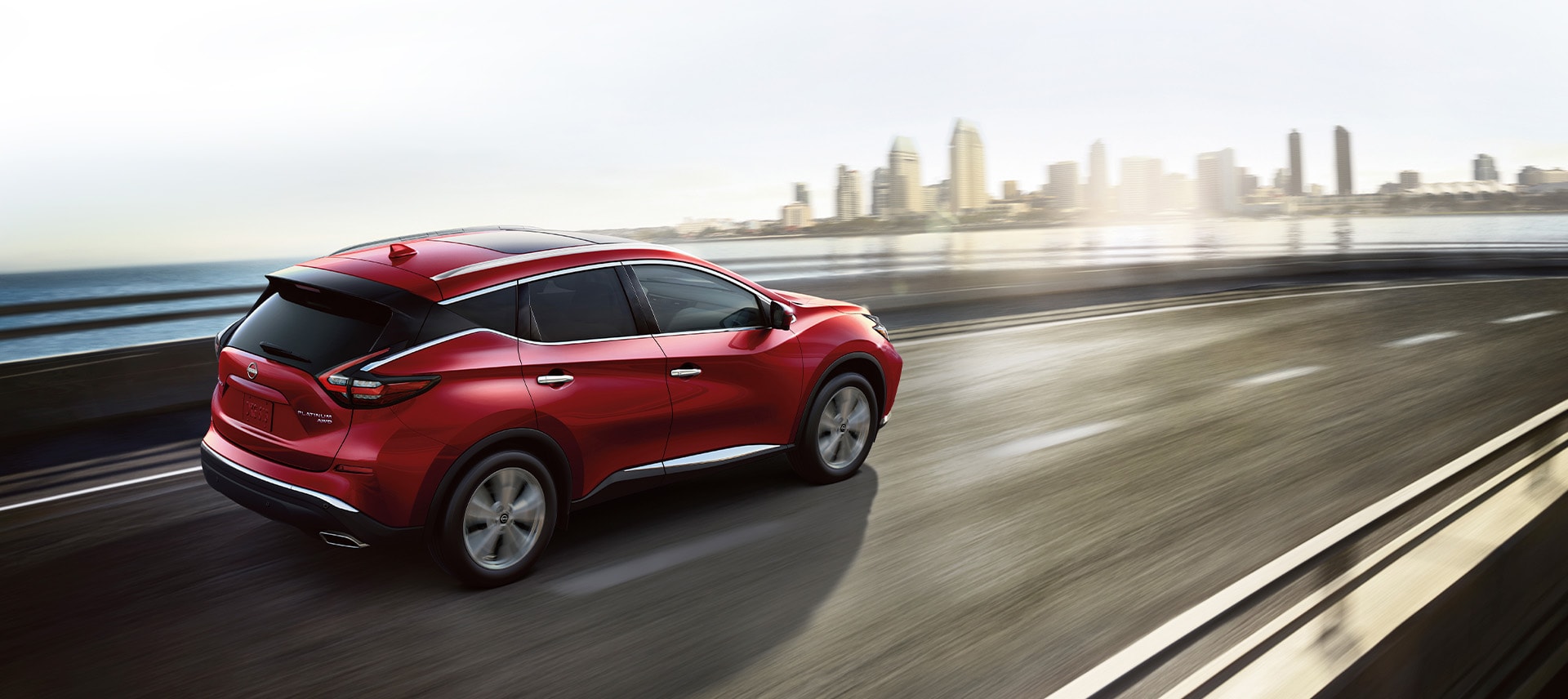 Red 2024 Nissan Murano driving fast on a bridge over water with a city in the background