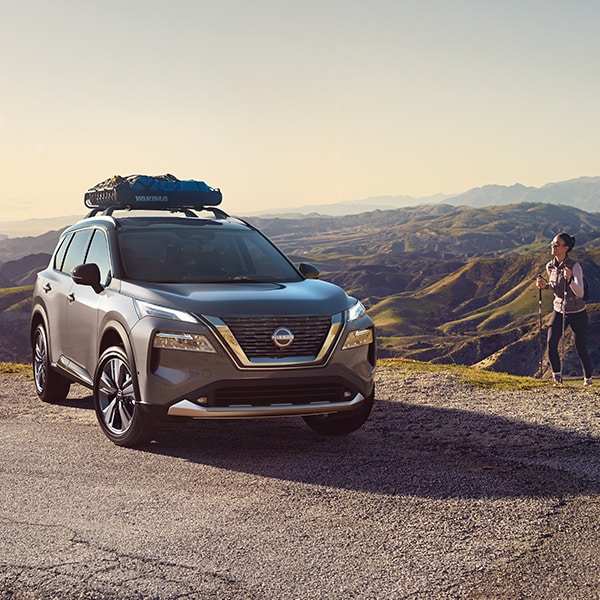 2024 Nissan Rogue with gear on the roof rails, on top of a hill, at a trailhead with hiker approaching