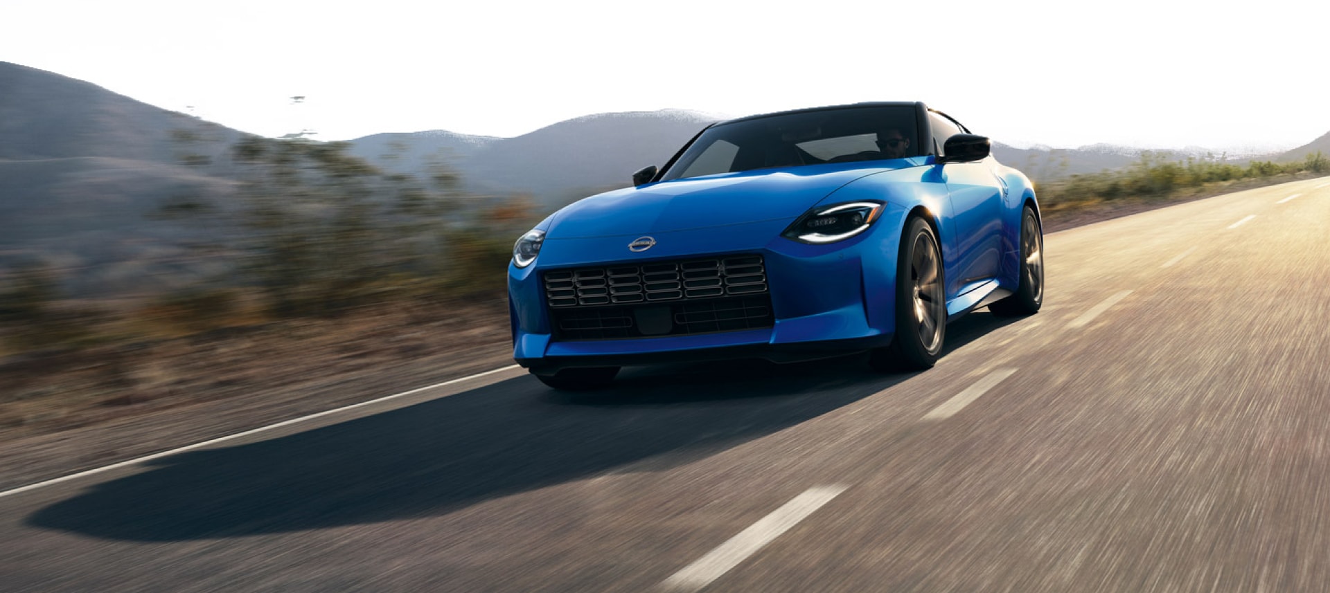 Angled front view of a blue 2024 Nissan Z driving fast on a road lined with brush